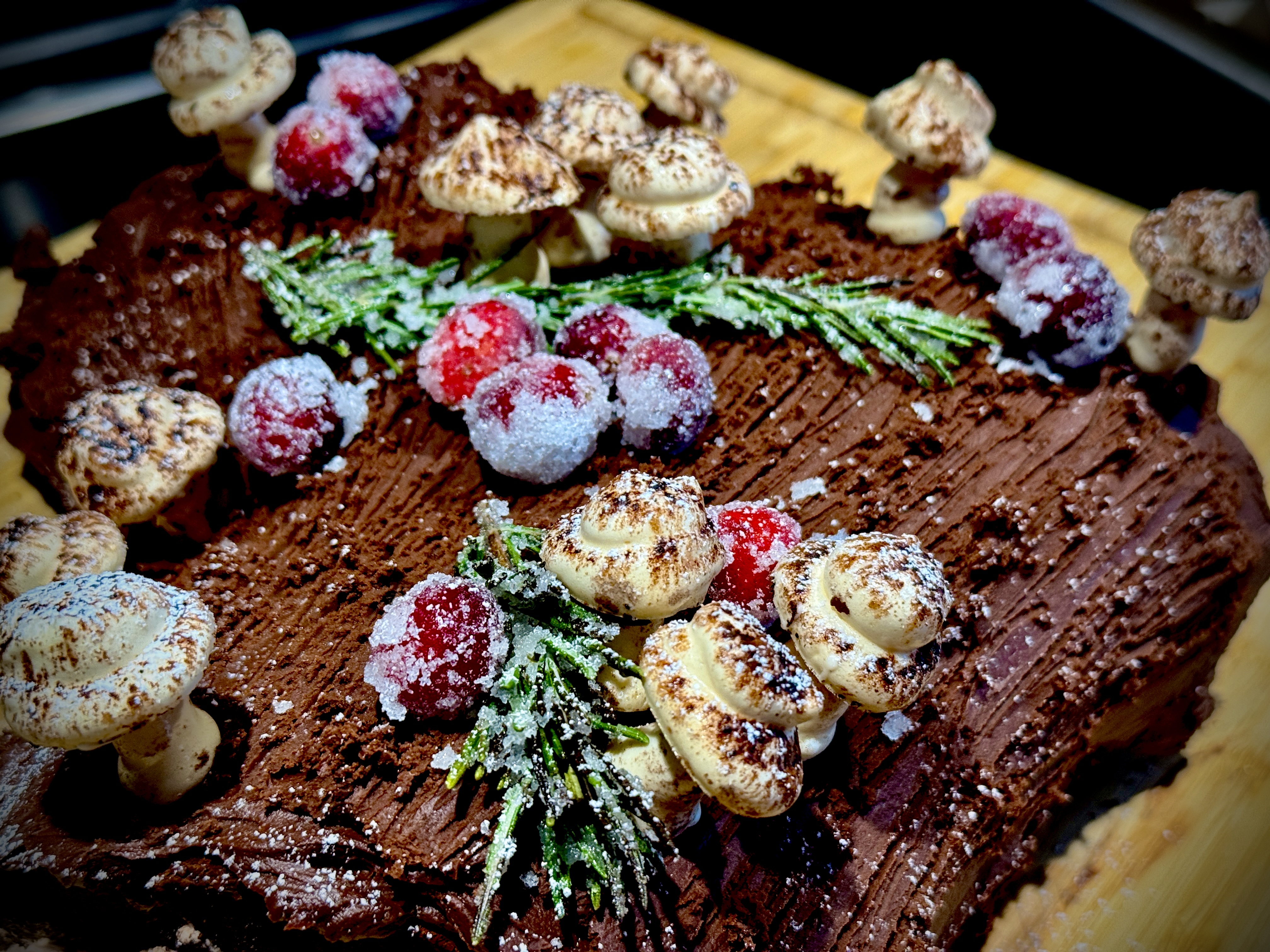 Close-up of decorated yule log with meringue mushrooms, sugared cranberries, and rosemary sprigs on chocolate ganache bark.