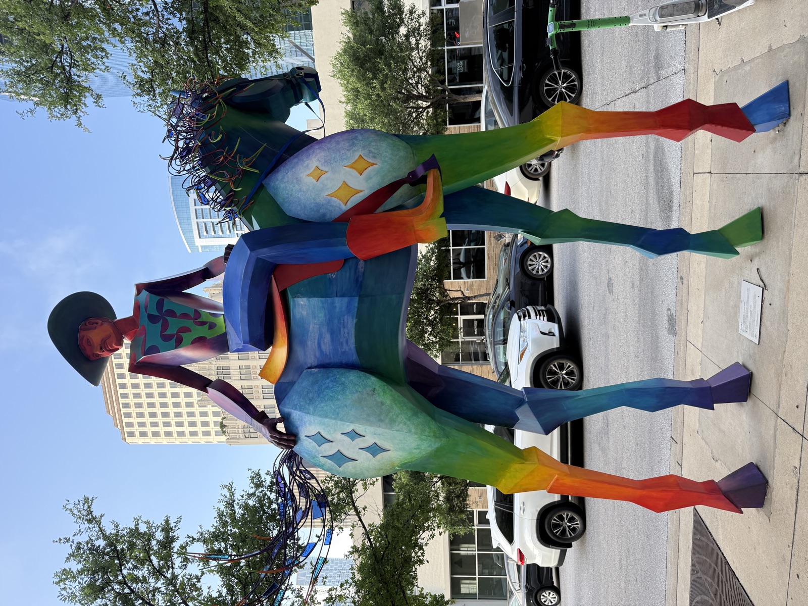 A monumental sculpture of a cowboy on horseback, maybe twelve feet tall, standing on a downtown Austin sidewalk. Both horse and rider are rendered in flat geometric panels painted in a rainbow gradient that runs from orange through yellow, green, blue, and purple along their bodies and legs. Pale starbursts decorate the horse&rsquo;s flanks. The horse&rsquo;s mane and tail are made of tangles of brightly colored translucent wire, exploding outward like fireworks. The rider wears a wide black hat and sits stiffly upright with one hand resting behind him on the horse&rsquo;s back. Behind the sculpture, parked cars and trees line the street, and several downtown office buildings rise into a clear blue sky.