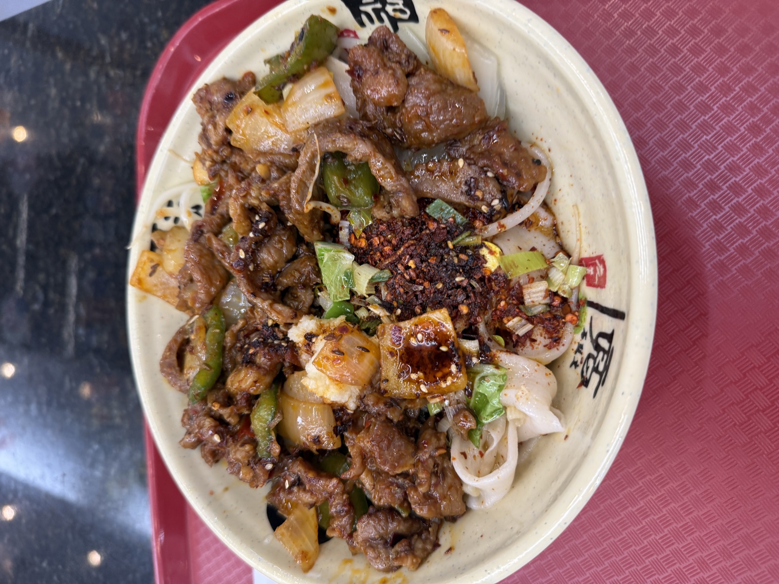 Overhead view of a generous bowl of biang biang noodles from Xi&rsquo;an Gourmet House, served on a red plastic tray. The wide, flat hand-pulled wheat noodles peek out from beneath an abundant topping of stir-fried lamb in a glossy dark sauce, mixed with chunks of white onion, sliced green bell pepper, and bright green leaves of leafy vegetable. A heaping pile of red chili flakes and toasted sesame seeds sits in the center, with a small drizzle of red chili oil pooled around a piece of onion. Scattered scallion greens add bright color throughout. The cream-colored ceramic bowl is stamped with red Chinese calligraphy characters.