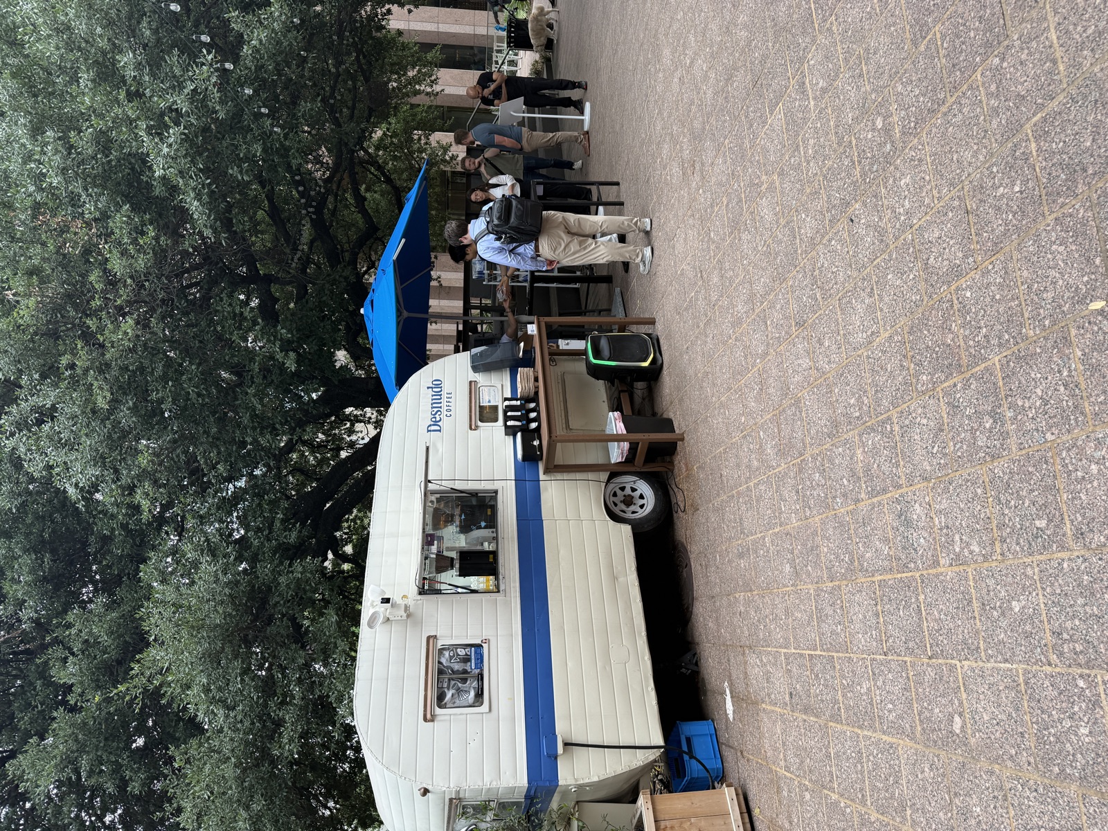 A small vintage white camper trailer with a horizontal blue stripe along its lower half and the word &lsquo;Desnudo Coffee&rsquo; painted in blue near the back, parked on a brick plaza beneath the spreading branches of a massive live oak. The serving window is open, with coffee equipment visible inside. A wooden side table holds three coffee dispensers, and a larger metal-topped service table sits next to the trailer with espresso machines and a tall blue umbrella overhead. A small line of customers stands waiting their turn, dressed in business-casual clothing. A string of cafe lights runs through the trees overhead, and a fluffy white dog can be seen at the right edge of the frame near the building.