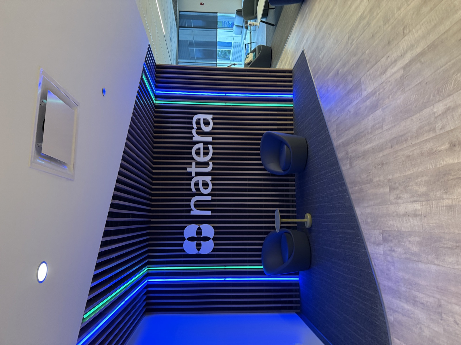 The Natera office lobby. The company name and logo, a clover-like cluster of four overlapping circles, are mounted in white on a feature wall made of vertical wooden slats. Two vertical strips of blue and green LED lighting flank either side of the wall, with the same colored light continuing across the slatted ceiling overhead. Two round blue swivel chairs sit on a dark gray rug in front of the wall, separated by a small mustard-yellow side table. Beyond the lobby, a glimpse of an adjacent room with more lounge seating and a window onto another office building.