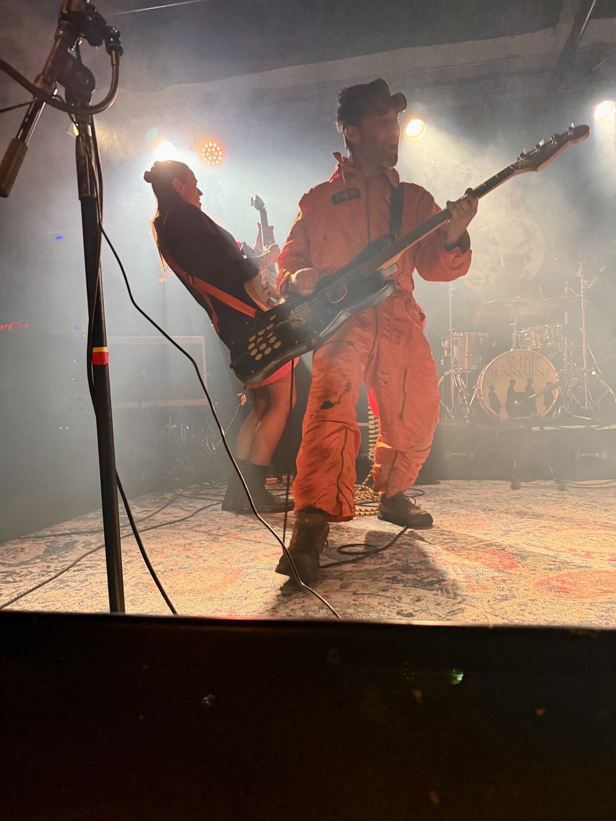 Coco in his orange flight suit playing bass in profile while bandmate Avona Nova leans back mid-performance, both lit by stage haze and colored light.