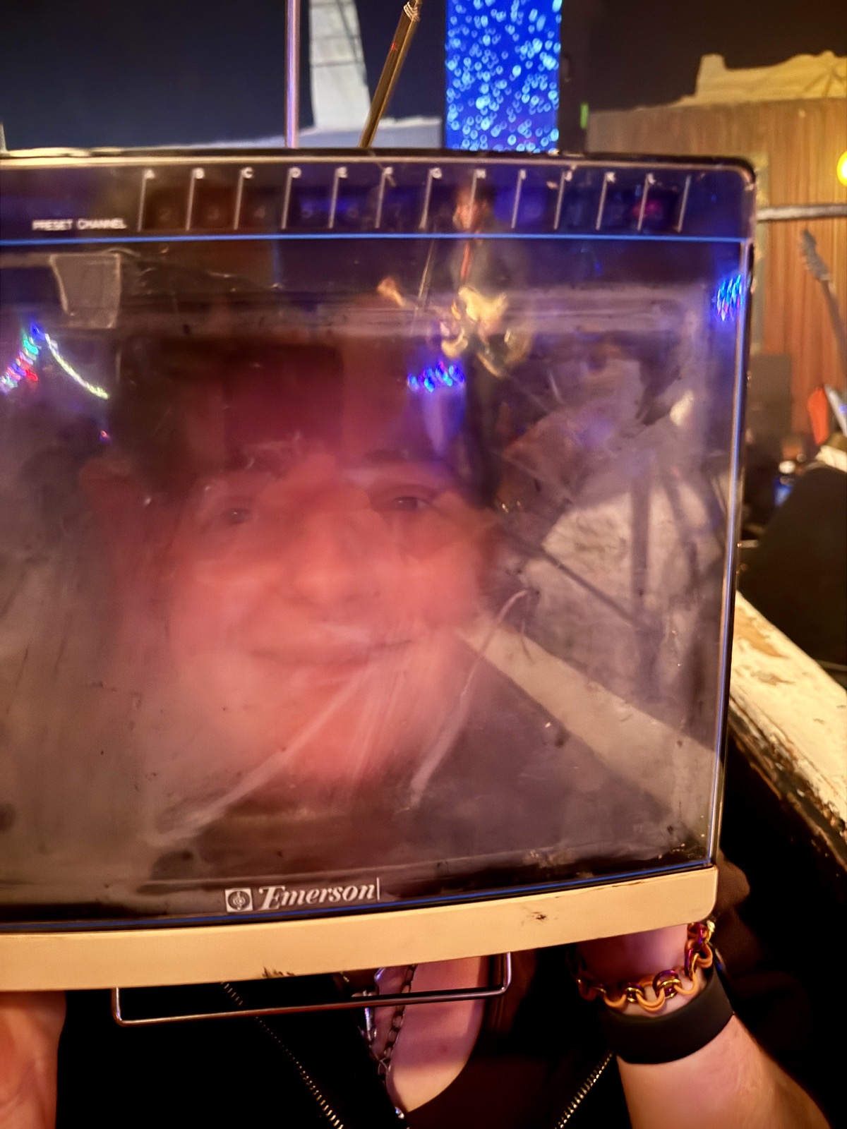 A face seen through the screen of a vintage Emerson television set worn as a helmet, lit pink and blue by the stage lights.