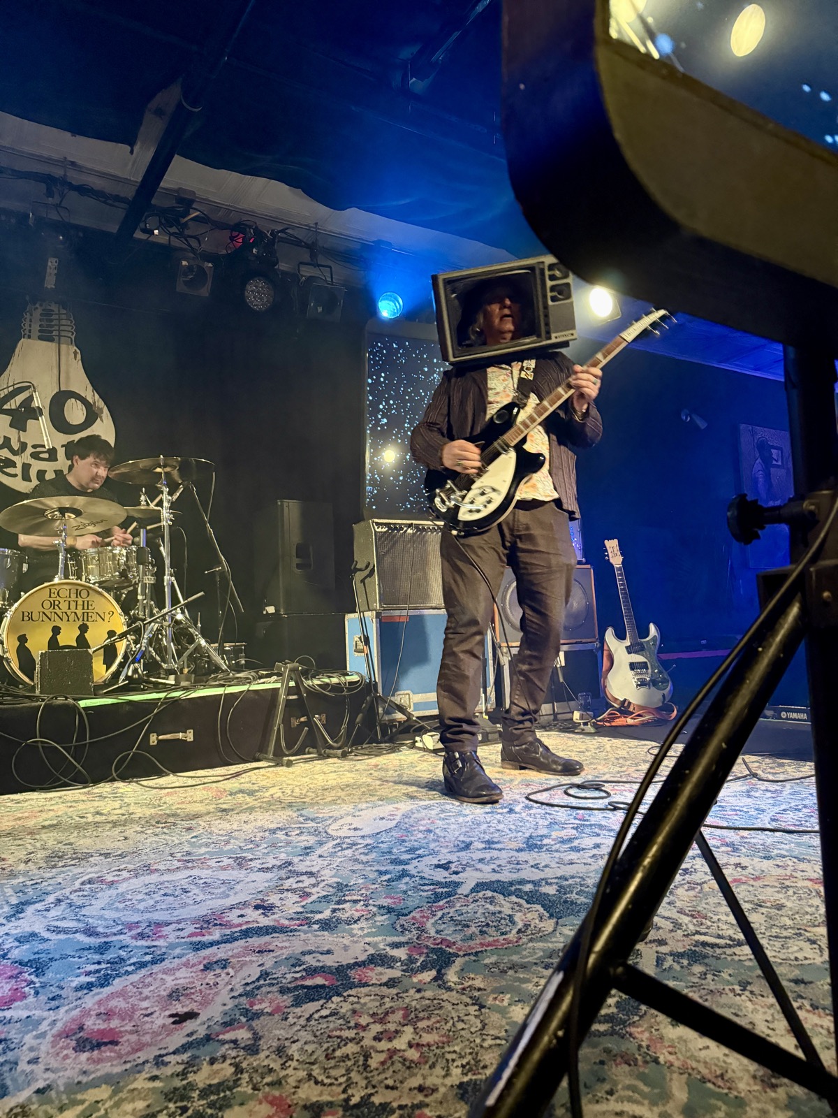 A guitarist wearing a vintage television set over his head plays a Rickenbacker on a rug-covered stage, drummer behind him and the 40 Watt light-bulb logo on the wall.