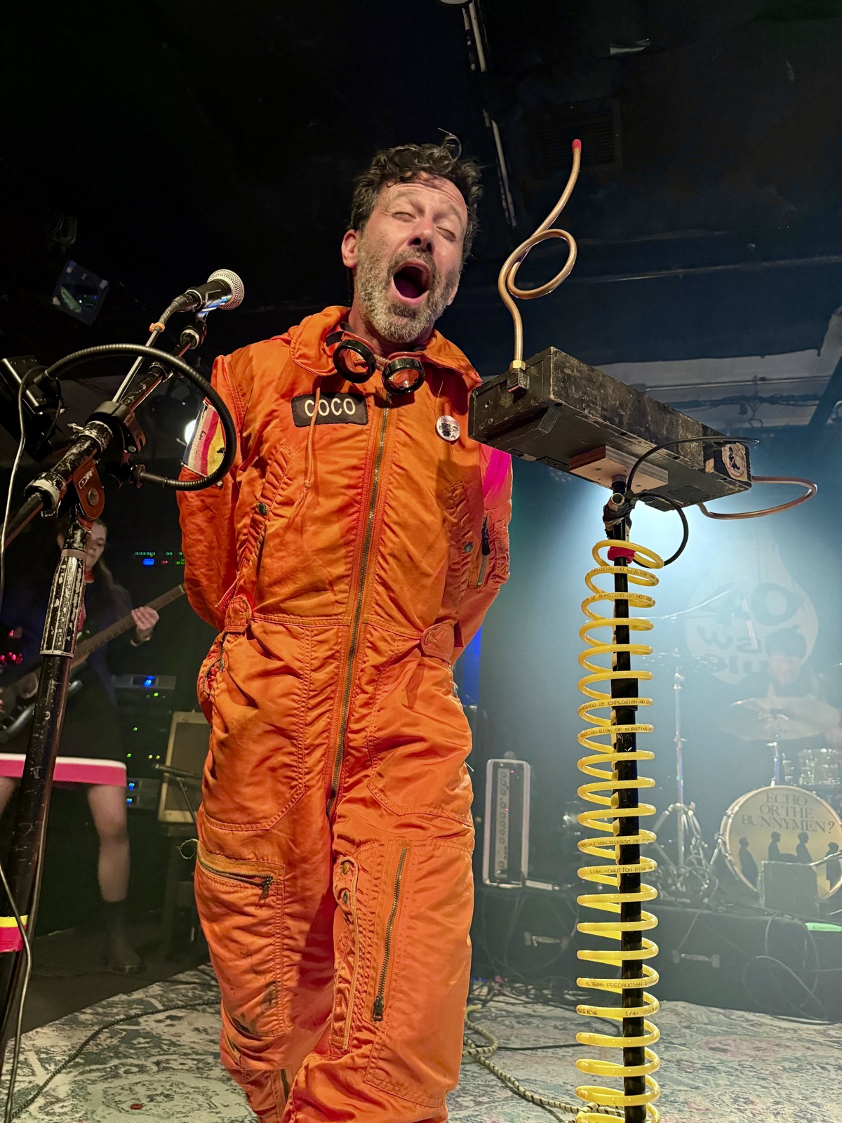 Coco of Man or Astro-man? in an orange flight suit with a COCO name patch and goggles, mouth wide open as he plays a theremin with his face beside a coil of yellow cable on stage at the 40 Watt.