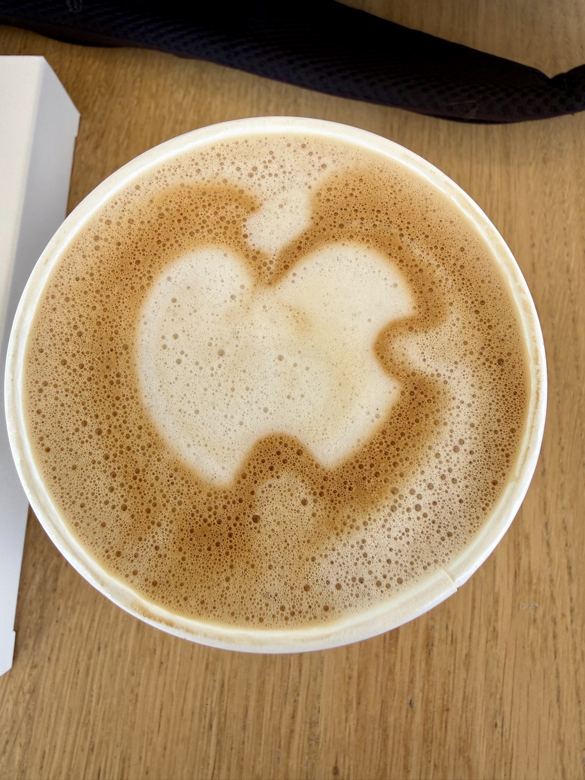 Looking straight down at a paper cup of latte on a wooden table. The foam and crema form two rounded lobes with a notch between them and a small stem at the top.