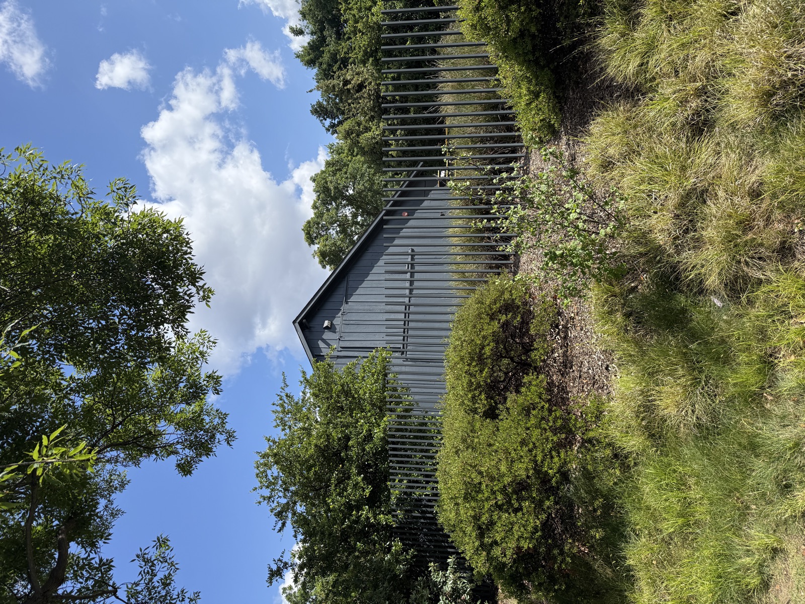 A dark gray board-and-batten barn with a steeply pitched roof, seen from below across a slope. A fence of tall, irregularly spaced gray slats stands in front of it, with shrubs and ornamental grasses around the base and a bright blue, partly cloudy sky above.