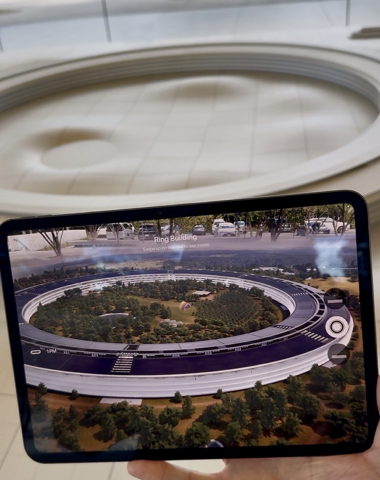 A hand holds a tablet at arm&rsquo;s length. Its screen shows a detailed 3D aerial render of a vast circular glass building ringed by trees, labeled &ldquo;Ring Building,&rdquo; with onscreen controls and a prompt to swipe up. Behind the tablet, blurred and out of focus, is a large white physical scale model of the same campus.