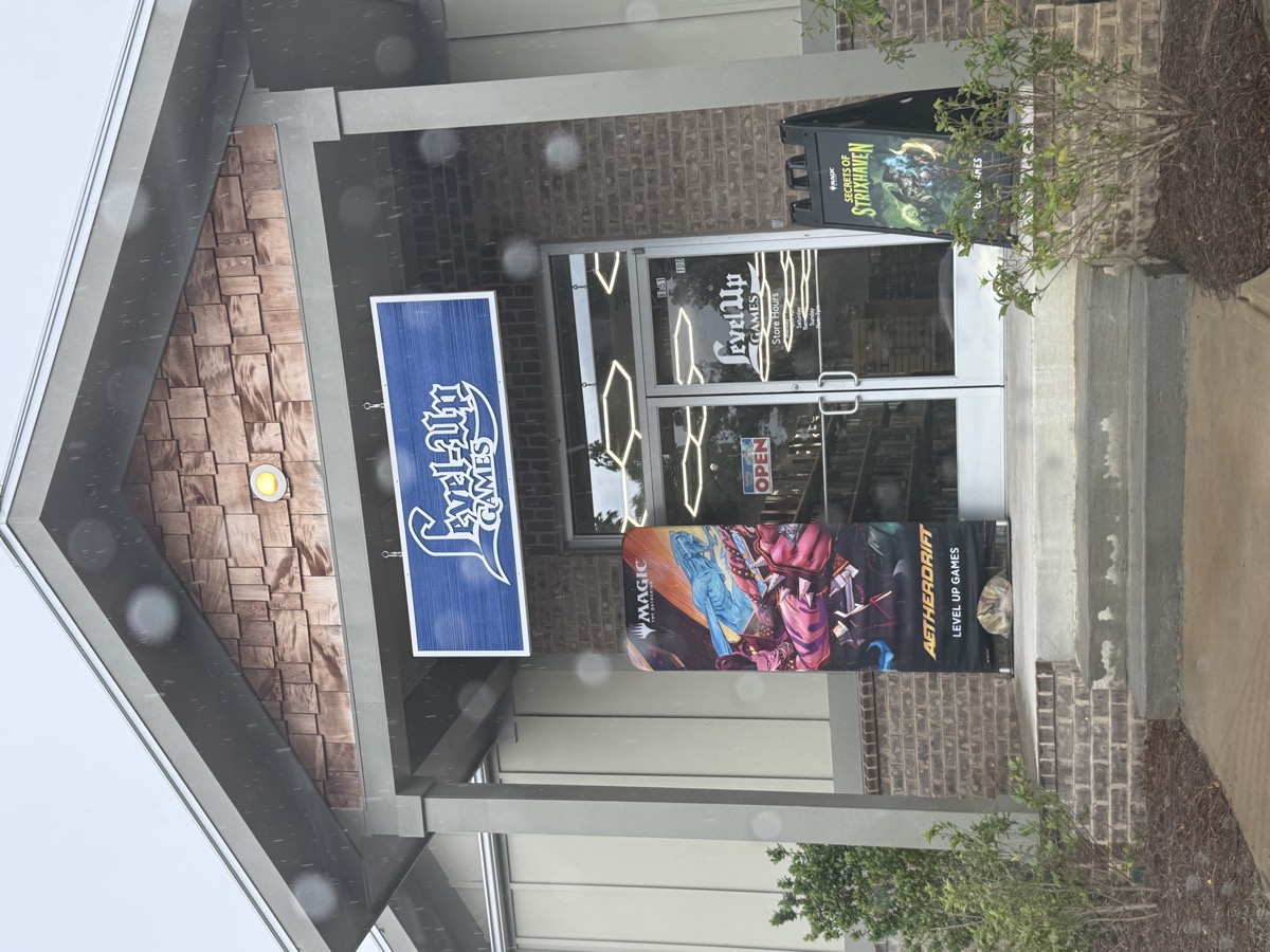 The covered brick entrance of a game store on a rainy day. A blue sign overhead reads &lsquo;Level-Up Games&rsquo; in white lettering, and two glass doors sit under hexagonal ceiling lights, flanked by tall Magic: The Gathering banners on the front steps. Rain spots speckle the lens.
