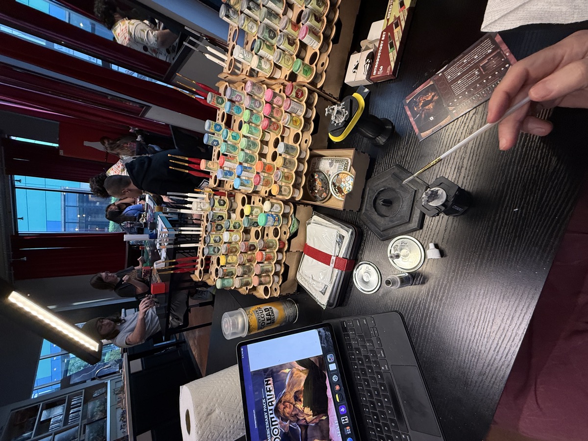 A painting table at a game store, seen from the painter&rsquo;s seat. An iPad propped on a keyboard case shows reference art for the Gloomhaven mercenary Hail. Tiered wooden racks hold dozens of small paint bottles, a can of Citadel spray primer, and rows of red-handled brushes. A primed miniature on a holder and a character card sit nearby, and a hand holds a fine brush in the foreground. Behind the table, other people paint and play beside tall windows with red curtains, dusk outside.
