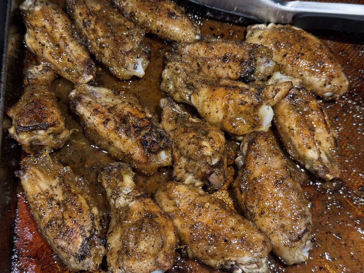 A sheet pan crowded with roasted chicken wings, browned and crisping at the edges, coated in dark herbs and spice and sitting in a shallow pool of their own rendered juices. A metal pan handle shows at the top of the frame.